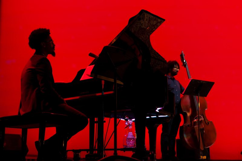Two musicians, one at a grand piano and one with a double bass, silhouetted against a vibrant red background.