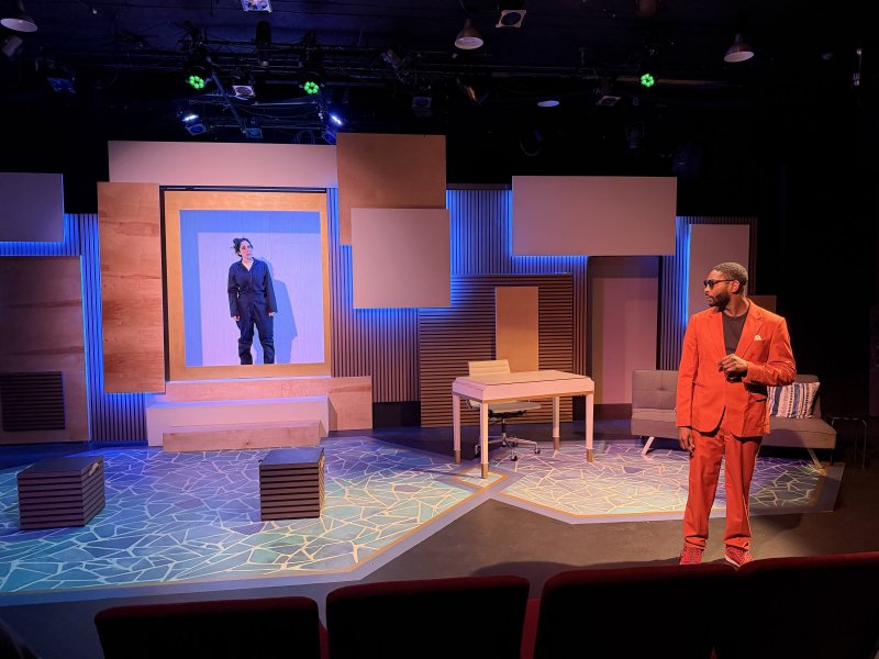 Man in red suit on stage with geometric floor, looking right. Woman in black jumpsuit on a screen, left. Blue lighting.