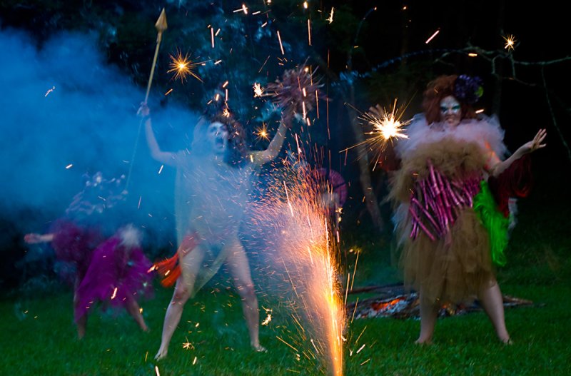 Outdoor performance art with costumed figures, sparklers, and smoke. One figure holds a spear, another wears a large brown and pink costume. A fire burns in the background.