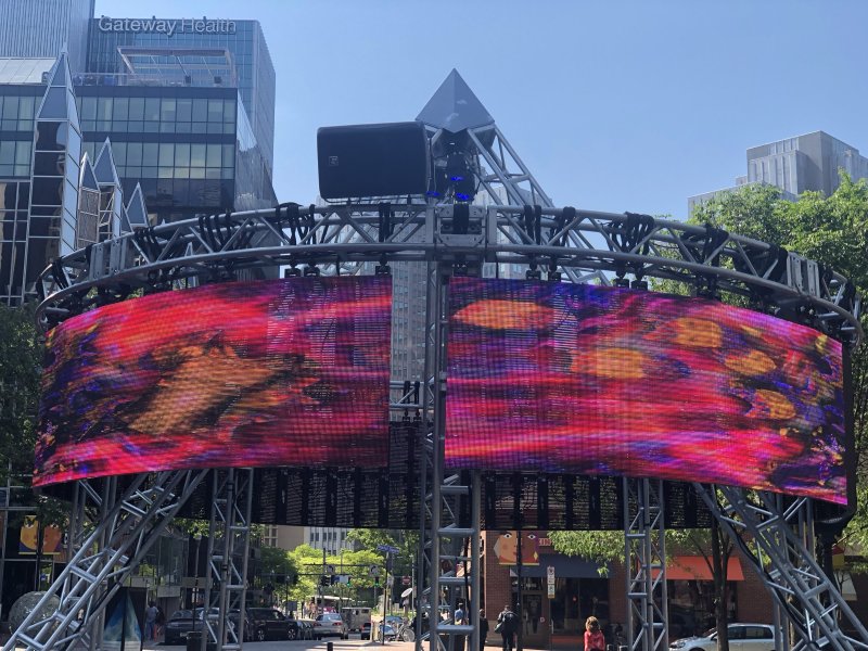 Large outdoor LED screen displays abstract red, orange, and purple patterns, framed by a silver metal structure.