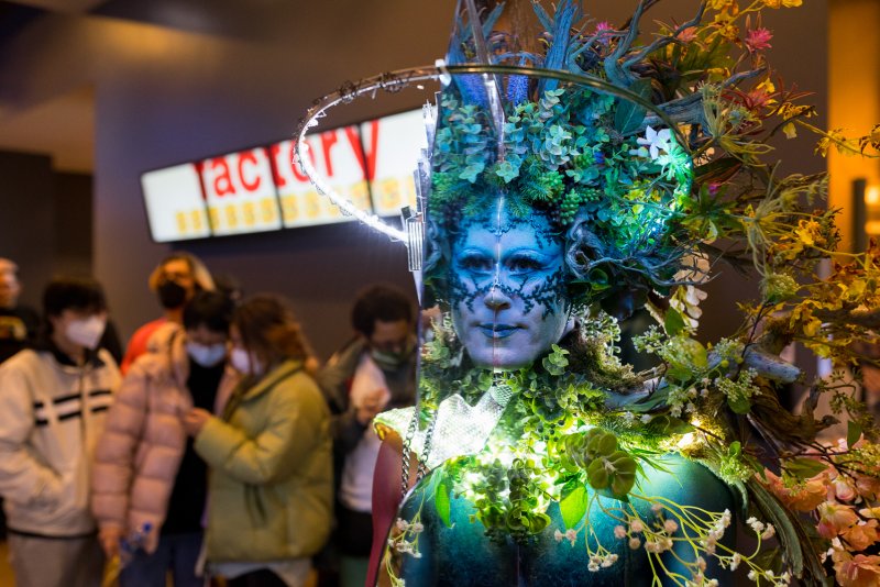 Performer in elaborate blue and green floral costume and makeup, with a glowing halo, stands in a crowded room.