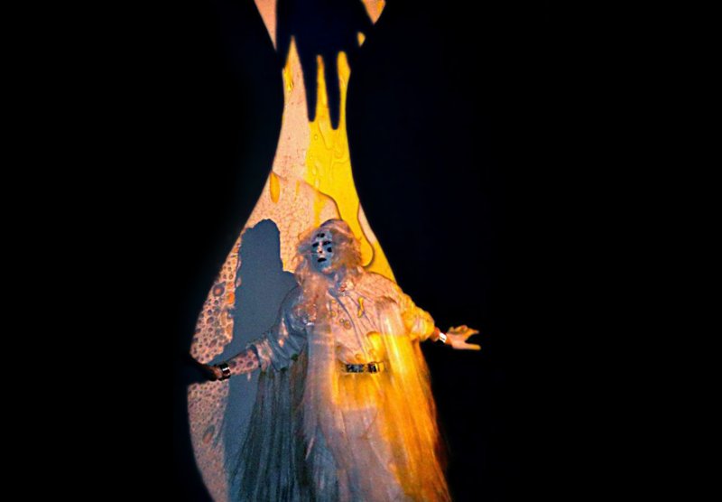 Figure in ornate costume with four eyes, centered in a black background. Figure is illuminated by yellow light and shadowed by a large hand.