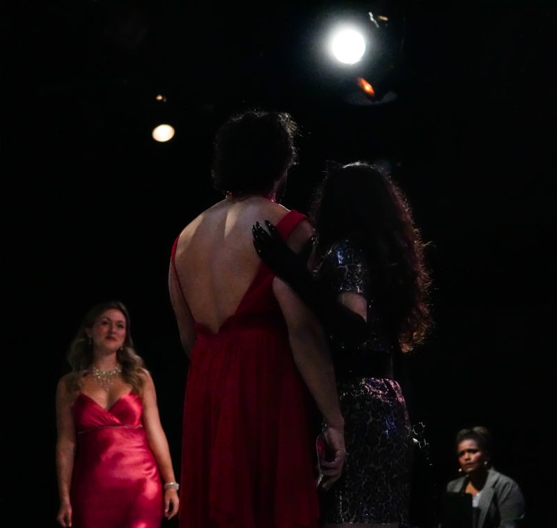 Two figures embrace on a dark stage, one in a backless red dress, the other in a sequined dress with a black glove.