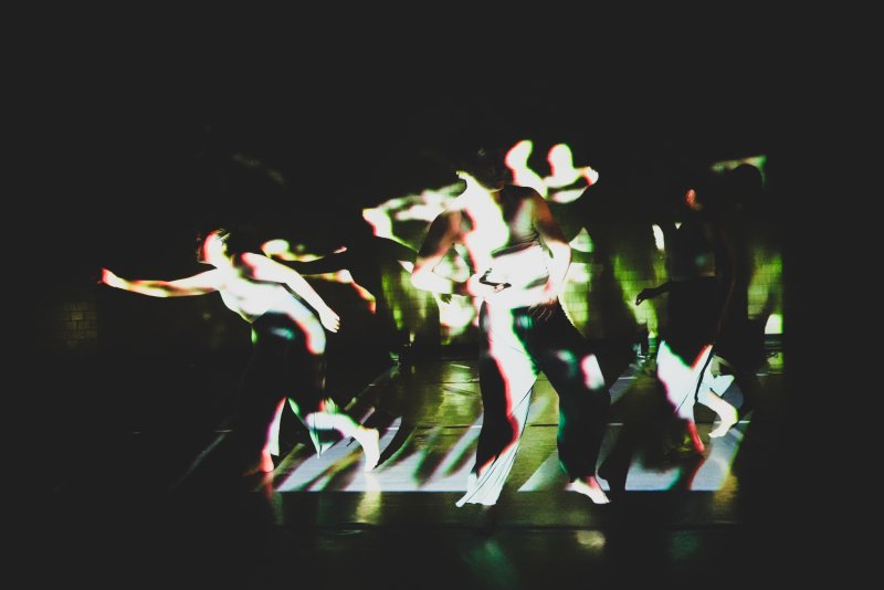 Three dancers in a dark space, illuminated by green and white light projections, creating blurred, ghostly figures.
