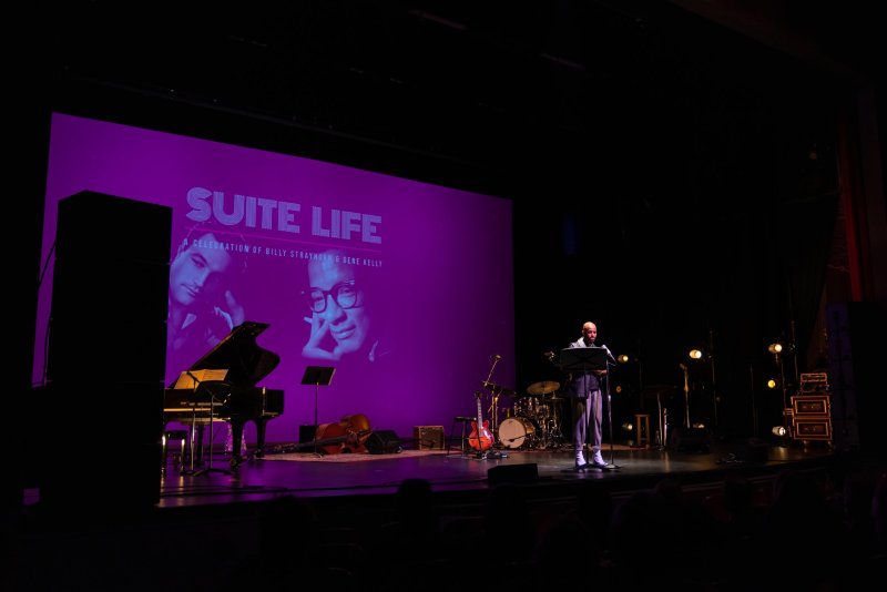A man reads from a podium on a stage with a grand piano, drums, and a large purple screen displaying "SUITE LIFE" and two men's faces.