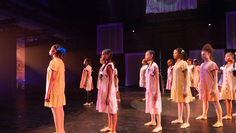 Stage view of young girls in light-colored dresses standing in rows, lit by purple and orange stage lights.