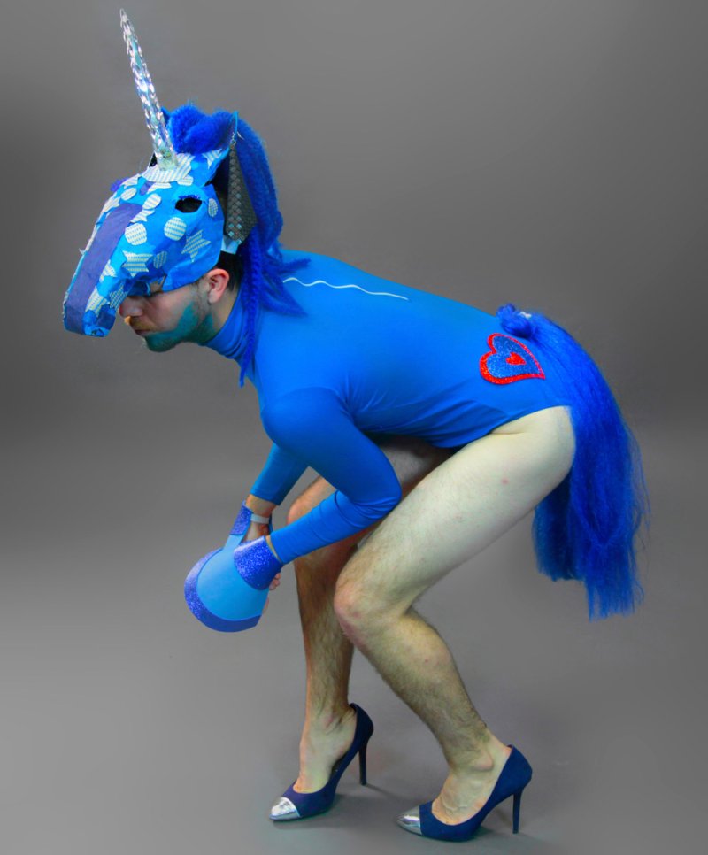 Man in blue unicorn costume, high heels, and blue body paint, bent over on a gray background.
