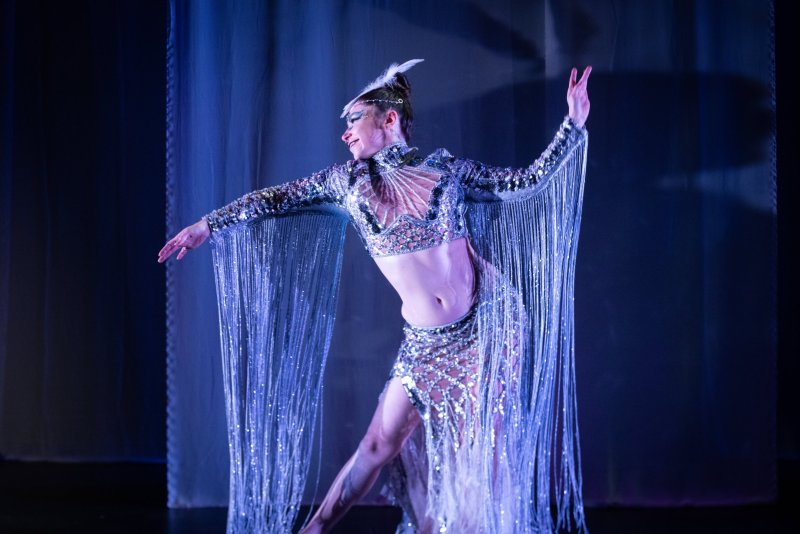 Dancer in a sparkling silver costume with long fringes, posing with arms outstretched against a dark blue background.