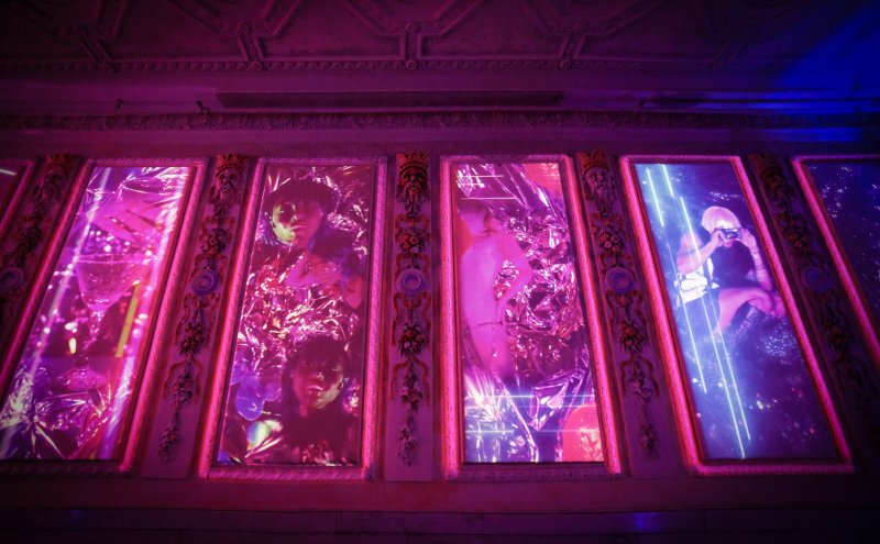 Five framed digital projections in pink and blue light show figures and objects, with crinkled plastic over some.