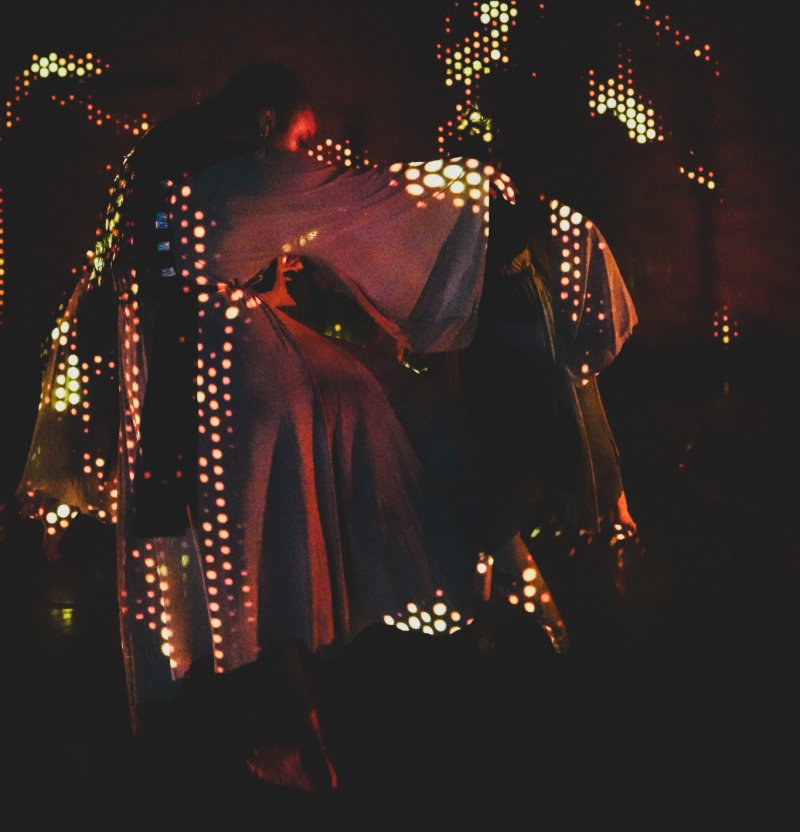 Two dancers in flowing garments, illuminated by warm, glowing digital patterns on a dark stage.