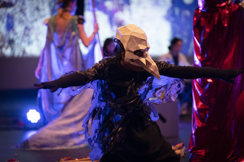 Performer in a white geometric bird mask and dark costume with outstretched arms, illuminated by blue light.