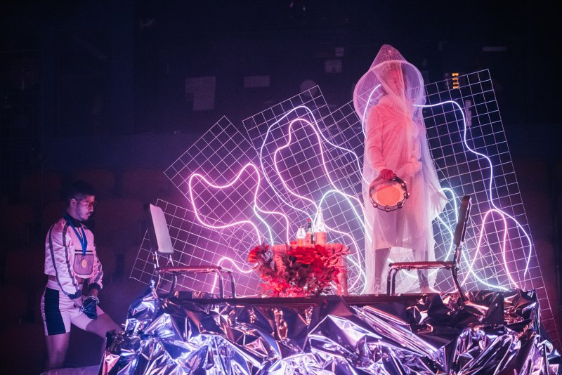 Performance with two figures, one in white translucent cloak holding a drum, other in white and black outfit, surrounded by neon grids.