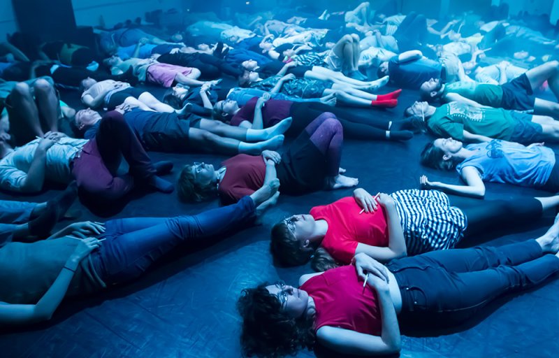People lying on a dark floor under blue light, some with eyes closed, wearing various casual clothes.