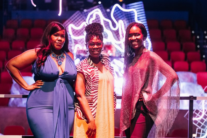Three Black women in vibrant outfits pose before a neon light sculpture in a red-lit room.