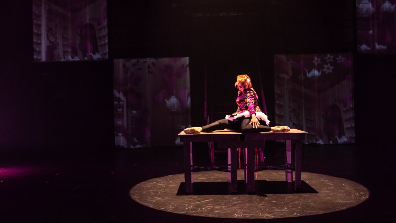Performance art piece. A dancer in floral jacket and black tights sits in a split on a table, lit by a spotlight. Behind, blurry projections of teapots and shelves appear on the dark stage.