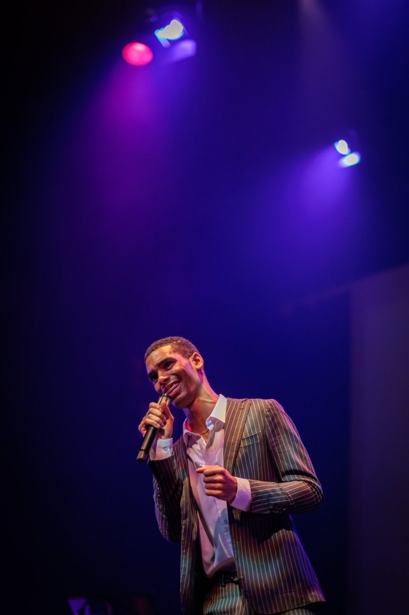 A man in a pinstripe suit sings into a microphone on a stage bathed in purple and blue spotlights.