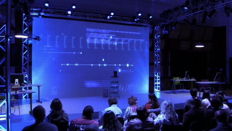 Audience watches a large screen displaying blue digital code and data visualizations in a dark room.