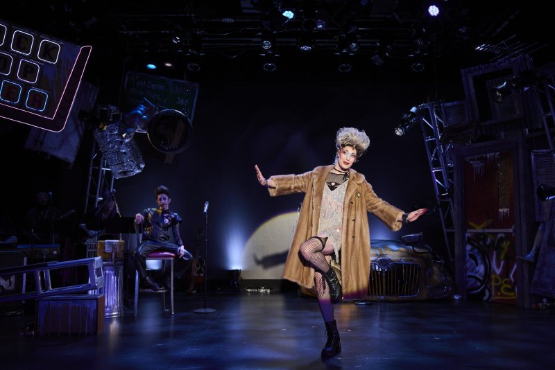 A drag queen in a fur coat, sequined dress, and fishnets poses on stage under bright lights, with a band member seated behind.