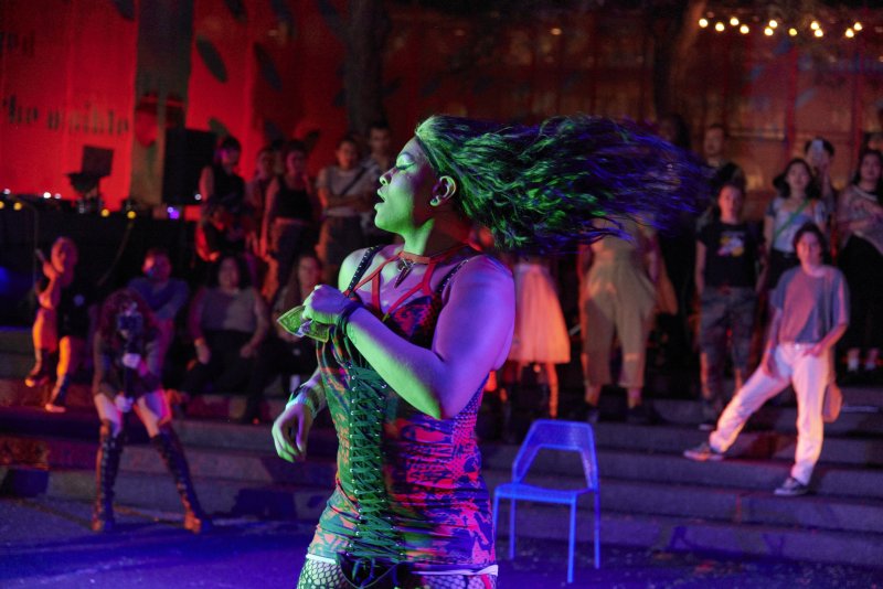 Outdoor performance scene with a person in a red and blue patterned corset and green hair, head thrown back, surrounded by an audience. Red light illuminates a backdrop, with string lights above.