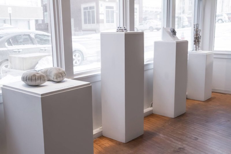 Art installation with four white pedestals, each displaying a unique sculpture, against a bright window backdrop.