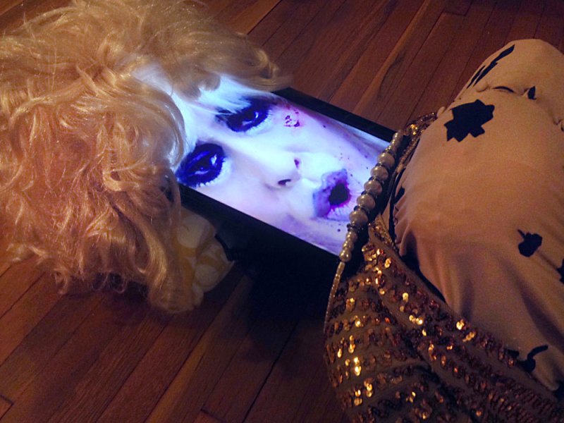 Blonde wig on a wooden floor, next to a screen displaying a pale face with dark eyes and purple lips.