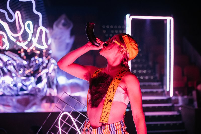 Person in orange hat and striped pants drinking from a horn, surrounded by neon lights and reflective surfaces in a dark room.