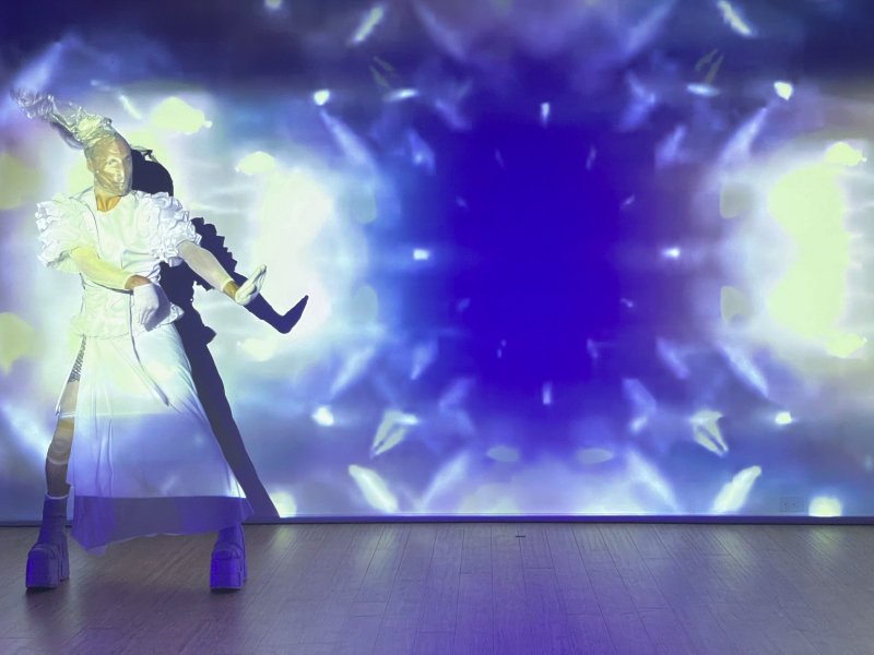 Performer in white costume, platform shoes, and elaborate headpiece striking a pose against a blue and white kaleidoscopic projection.