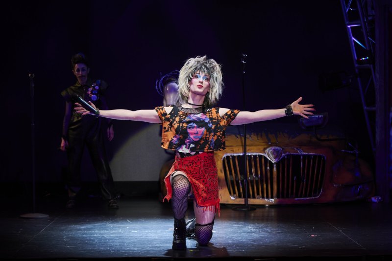 Performer in blonde wig, crop top, fishnets, and red skirt kneels center stage, arms outstretched, holding a microphone.