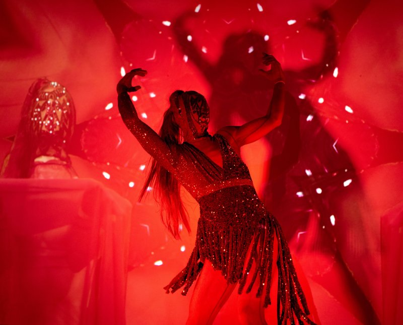 Performer in a shimmering, fringed costume and elaborate mask, arms raised, dancing on a red-lit stage with light reflections.