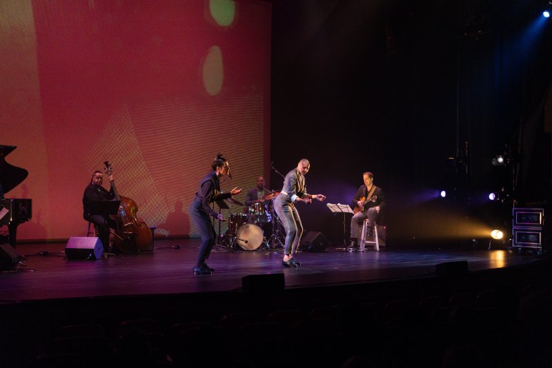 Two dancers in dark suits perform on a stage with a band, bathed in red and purple stage lights.