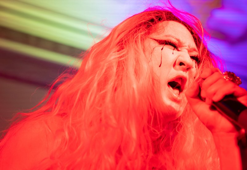 Close-up of a person singing into a microphone, bathed in red light. Black streaks run from their closed eyes. Background is a blur of green and purple.