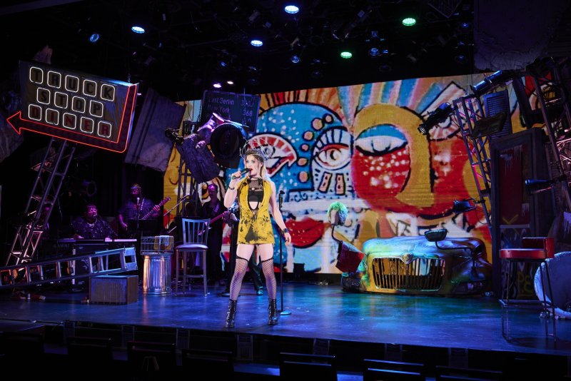 Performer in yellow dress and fishnets sings on stage with a band, backed by a colorful, graffiti-style mural and industrial set pieces.