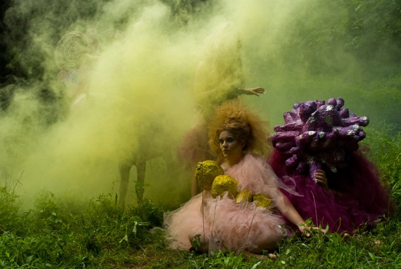 Outdoor scene with figures in elaborate costumes amidst green grass and yellow smoke. One figure wears a pink tulle dress with yellow objects, another a purple coral-like structure.