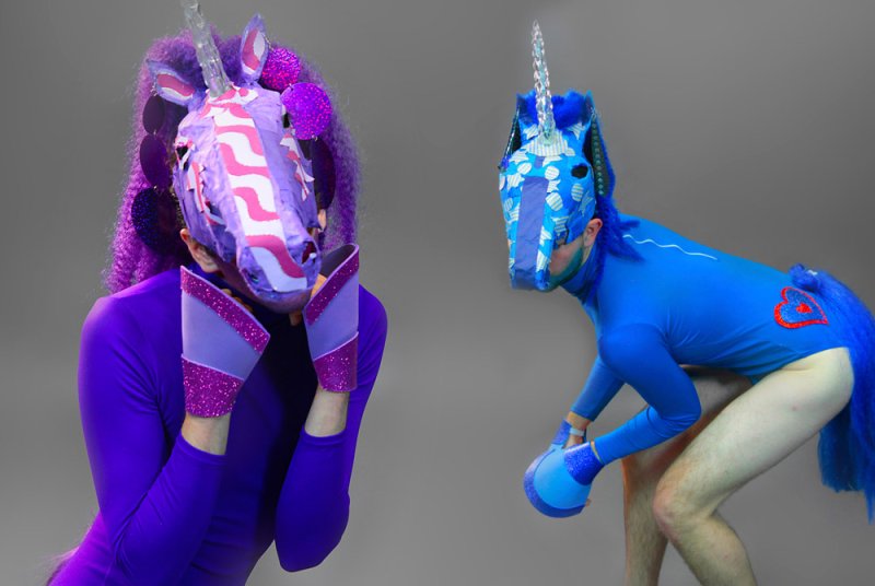 Two people in full-body unicorn costumes, one purple and one blue, with glitter accents on a gray background.