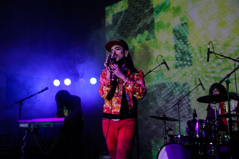 Performer in a colorful jacket and red pants singing into a mic on stage, with a keyboardist, drummer, and green projections.