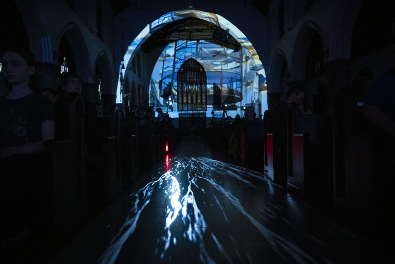 Dark church interior with blue light projected onto the arched ceiling and floor, creating abstract patterns and illuminating shadowy figures.