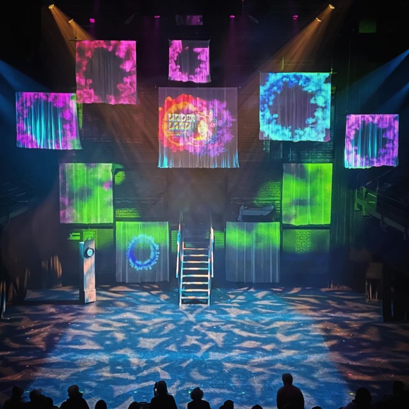 Theater stage view, lit with colorful projections on hanging fabric squares. Stage floor is patterned with blue light, with stairs leading to a platform. Audience silhouettes in foreground.