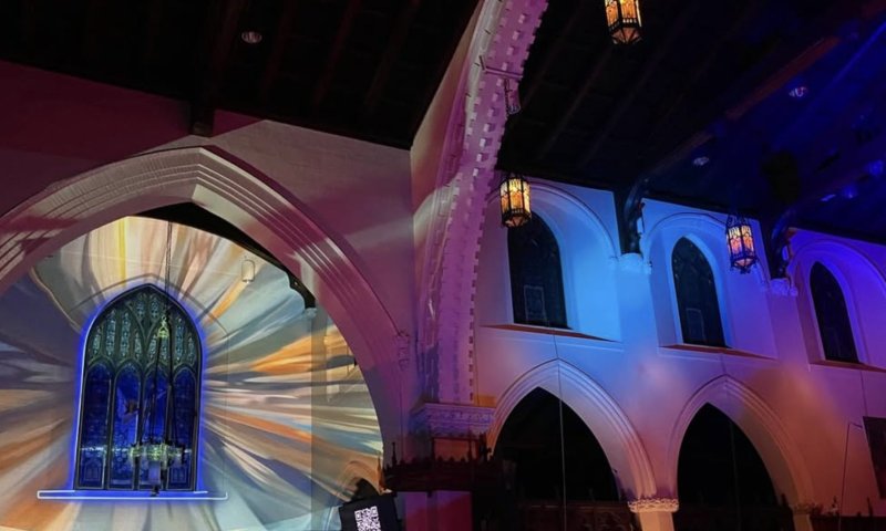 Church interior with colorful light projections on white walls, arched windows, and hanging lanterns.