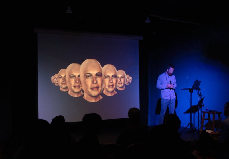 Darkly lit stage with a presenter standing near a laptop. A projection screen displays a row of bald male heads. Silhouetted audience in foreground. Blue lighting on stage right.