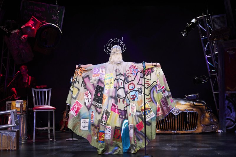 Blonde performer in a white wig and crown, wearing a long, colorful cape adorned with punk rock posters, facing away from the viewer on a stage.