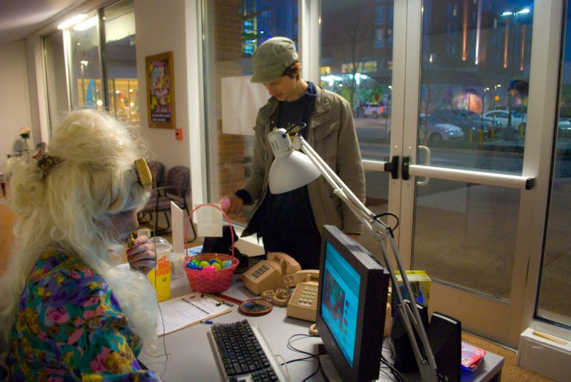 A person in a blonde wig and floral top sits at a desk with a monitor, while a man in a cap stands nearby.
