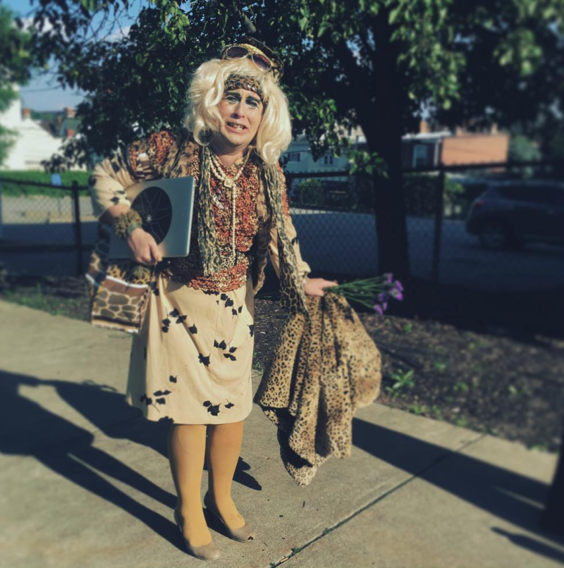 Blonde drag queen in leopard print and beige dress holding a laptop and flowers on a sunny sidewalk.