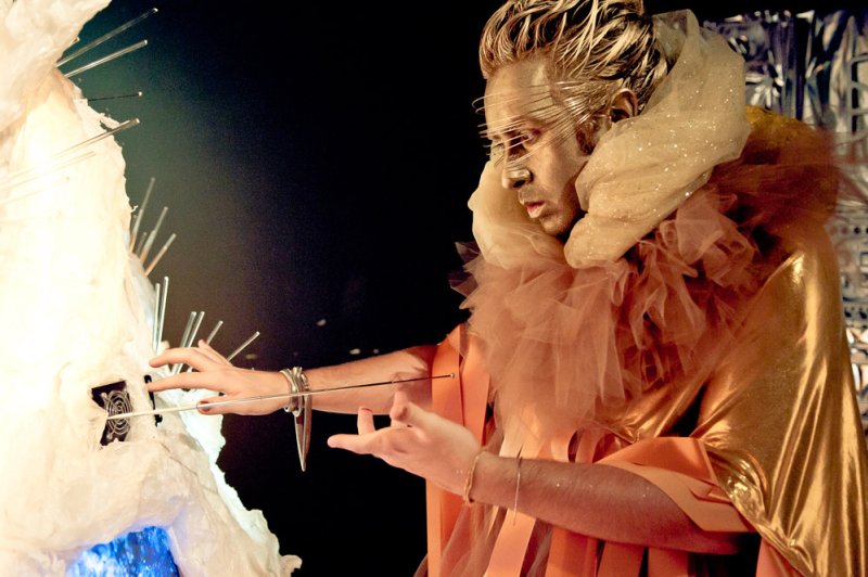 Performer in gold makeup and costume with ruffled collar, touching a white, spiky sculpture with blue light.