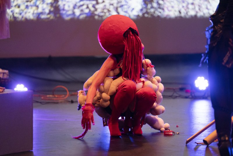 Performance art featuring a figure with a large red spherical head and red dreadlocks, crouching amidst a cluster of soft, fleshy-toned forms. The figure wears red pants and has a red hand extended towards the floor. The background is a blurry stage 