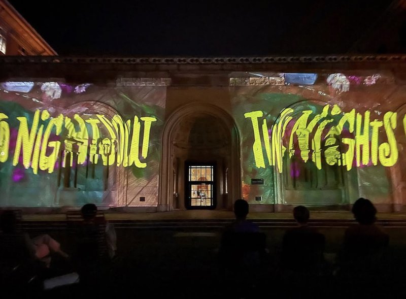 Nighttime projection of "GOOD NIGHT" and "SWEET DREAMS" in yellow graffiti-style text on a stone building, with green and purple washes.