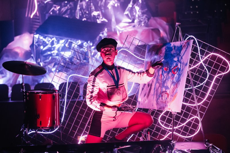 Performer in beret and white outfit painting a blue and red abstract face on an easel, surrounded by neon grids and drums.