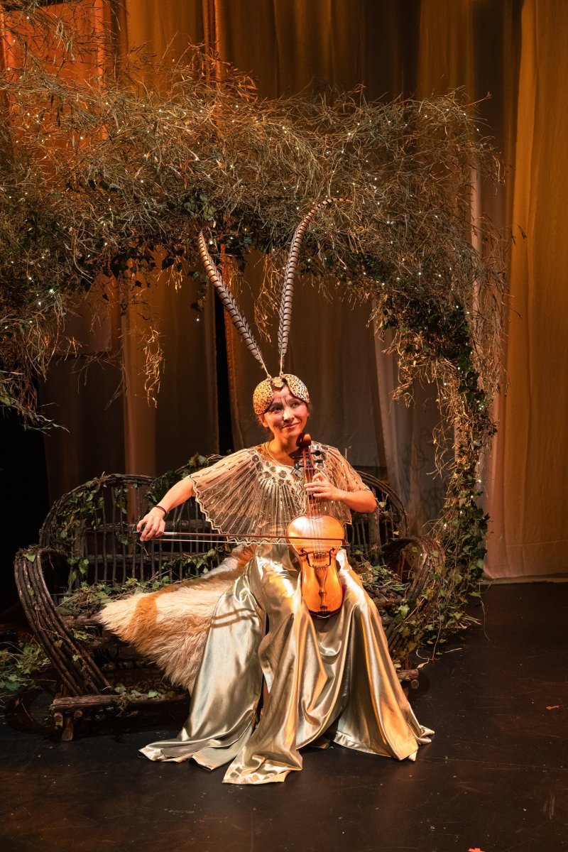 Woman in gold dress and feathered headpiece playing violin on a rustic bench under a leafy arch with twinkling lights.