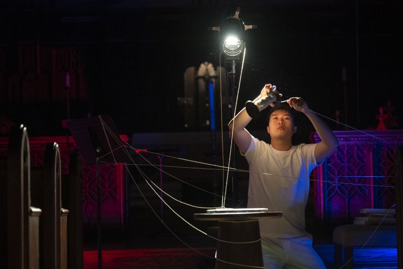 A person in a white shirt adjusting strings connected to a spotlight, with red and blue light on dark church pews.