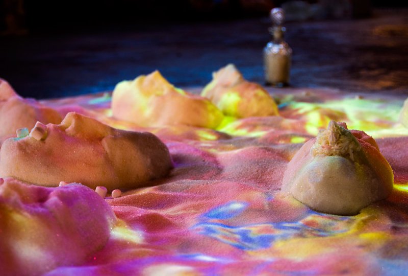 Close-up of a sand installation with small mounds, some topped with white objects, illuminated by colorful lights.