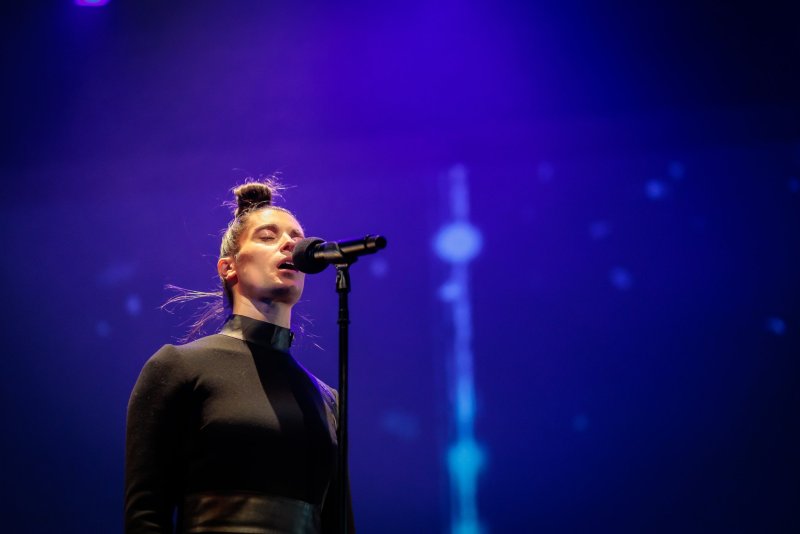 Woman with hair in a bun singing into a microphone on stage, illuminated by purple and blue lights.